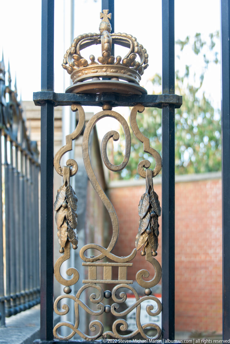 Gilded crowned monogram and scrolling ornament set into a black iron gate.