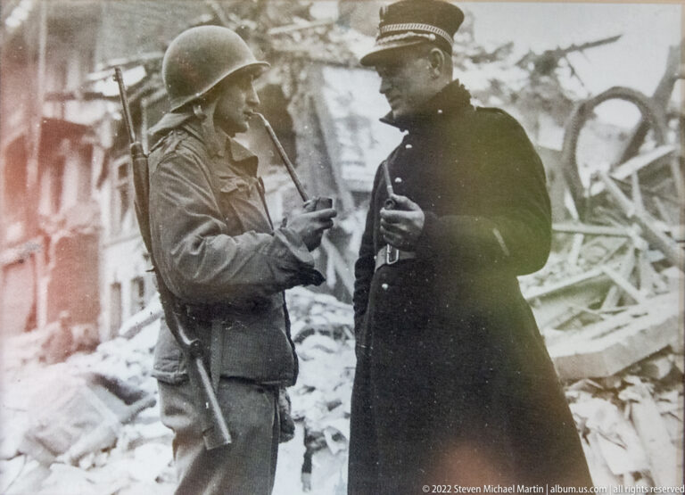 Black-and-white wartime photograph of a U.S. soldier and uniformed officer speaking amid heavily damaged buildings and rubble.