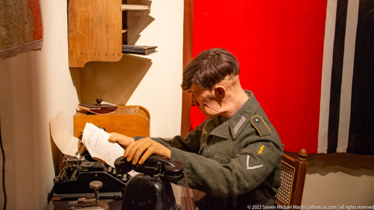 Uniformed seated operator typing on a typewriter in a reconstructed wartime communications or administrative room.