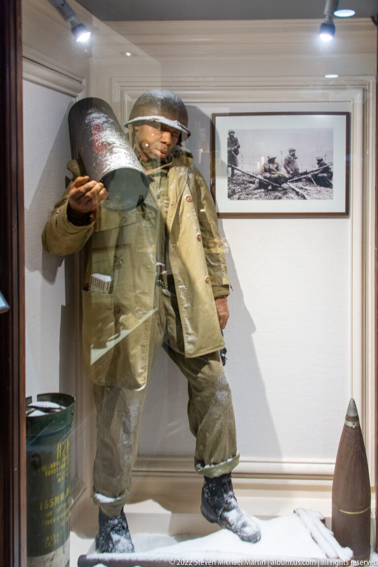 Two helmeted U.S. soldiers carry a large cylindrical container across a snow-covered museum diorama beside a large projectile.