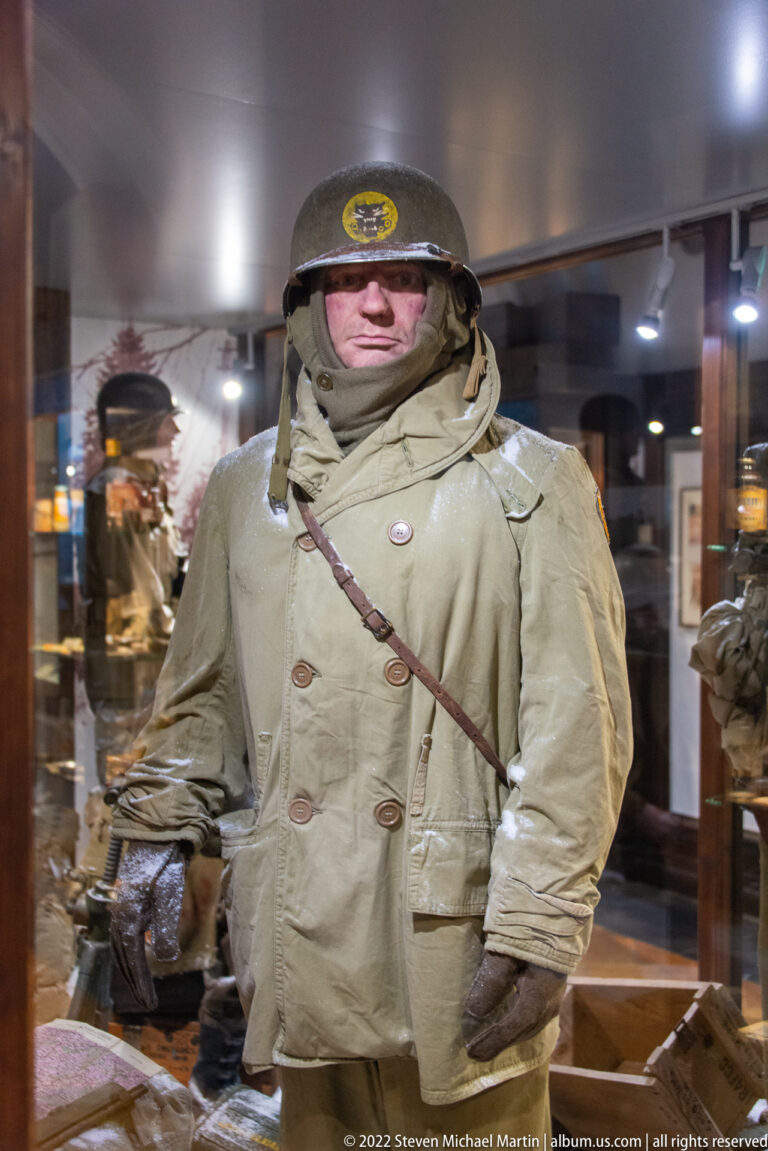U.S. soldier mannequin in heavy winter overcoat, steel helmet, gloves, scarf, and shoulder-slung field gear.