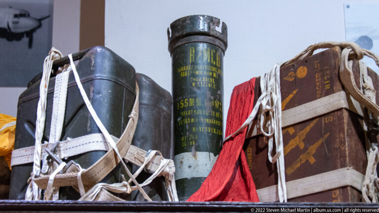 Close display of tall cylindrical supply containers, white straps, cords, and bundled airborne equipment with stenciled markings.