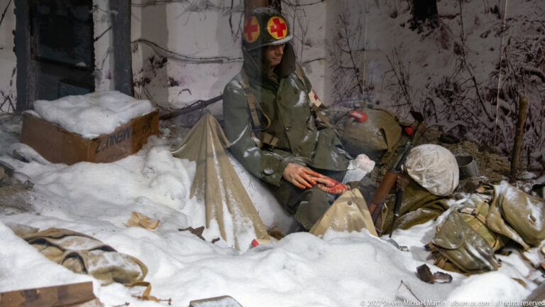 Combat medic with Red Cross markings tending a wounded soldier amid snow, packs, crates, and field equipment.