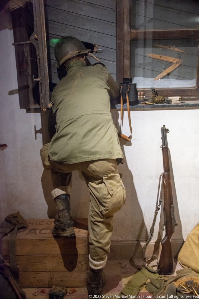 Helmeted U.S. soldier crouching at a dark window opening with a rifle propped against the wall nearby.