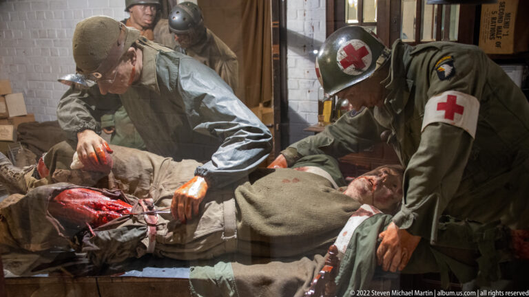 Two medics with Red Cross markings treat a badly wounded soldier in a graphic wartime aid-station diorama.