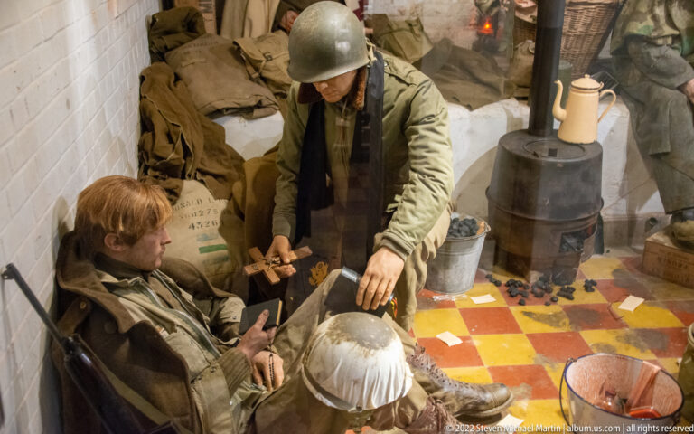 Two U.S. soldiers in cramped quarters beside a small stove, kettle, rifle, sacks, and red-yellow tiled floor.