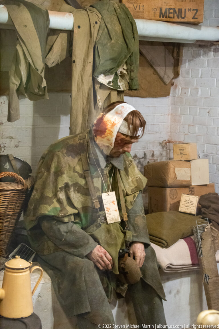 Bandaged wounded soldier seated among blankets, clothing, supply boxes, and an enamel vessel in a field-care display.