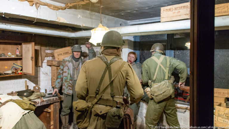 Several soldiers stand in a cluttered low-ceilinged room with crates, clothing, equipment, exposed brick, and overhead pipes.