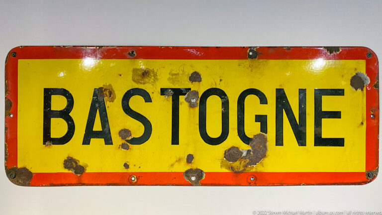 Weathered yellow road sign with red border reading BASTOGNE, marked by numerous punctures, chips, and dark impacts.