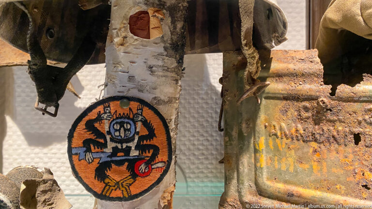 Round embroidered insignia with orange and black figure displayed beside heavily rusted metal military relics.