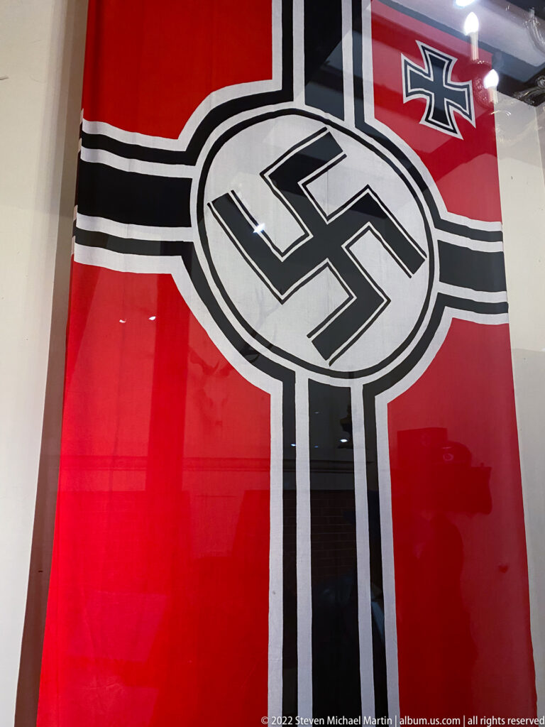 Large red Nazi flag with central swastika, black-and-white cross design, and small Iron Cross-style emblem behind glass.