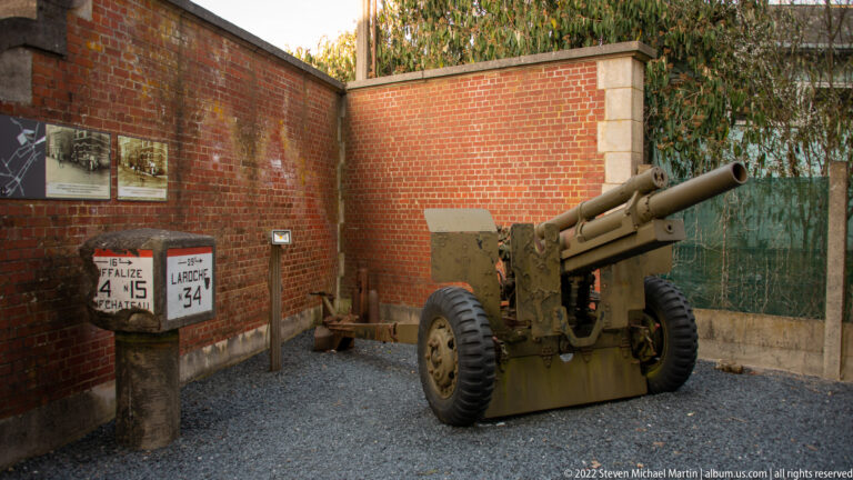 Towed field gun with large wheels displayed on gravel beside brick walls and wartime road signs.