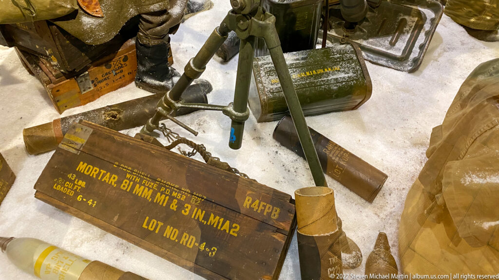 Wooden crate stenciled for 81 mm mortar ammunition among cylindrical tubes, metal containers, and field equipment.
