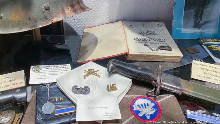 Display case with signed unit book, handgun, cloth patches, medals, badges, and labeled personal veteran memorabilia.