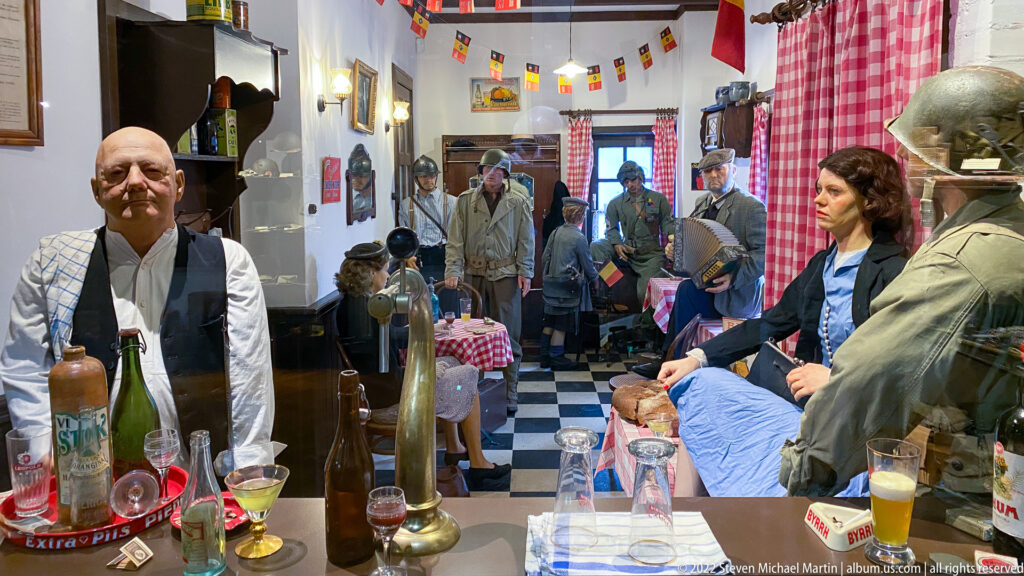 Reconstructed café interior with bottles, glasses, civilians, and uniformed soldiers beneath small flags and checked curtains.