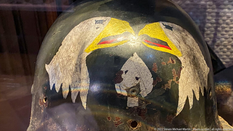 Dark steel combat helmet with large painted white wings, yellow-red details, holes, and heavy surface wear.