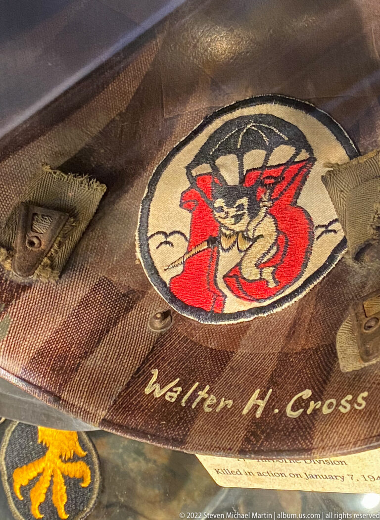 Close-up of worn military textile with circular red cartoon-style emblem, the name “Walter H. Cross,” and nearby insignia.