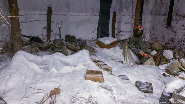 Uniformed soldier in a snowy woodland diorama surrounded by trees, packs, crates, helmets, and scattered field equipment.