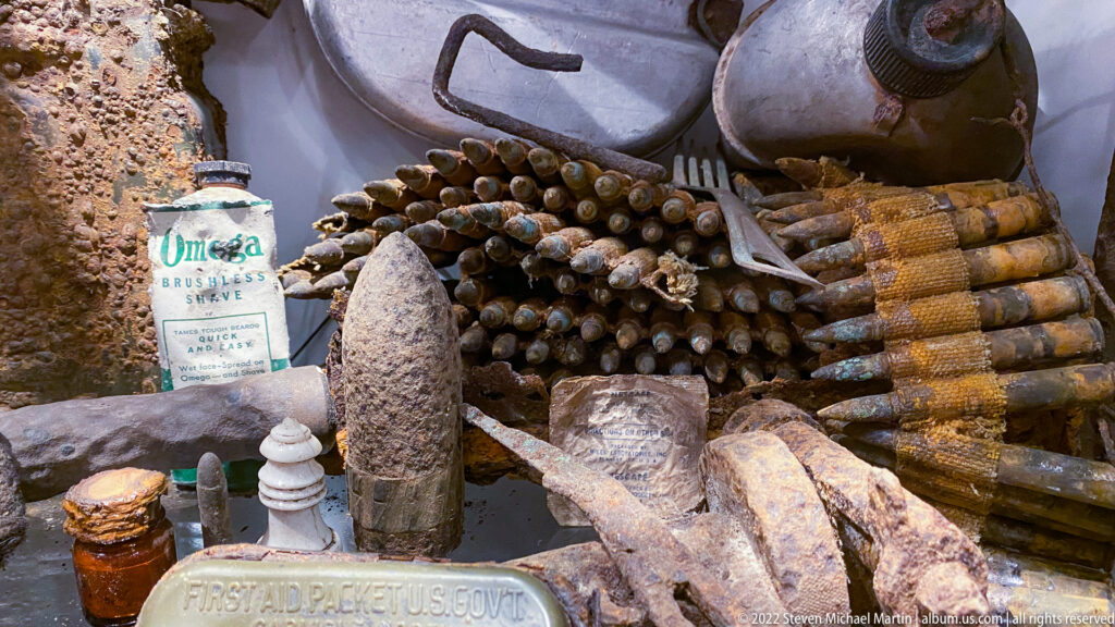 Corroded ammunition, bottles, metal fittings, ceramic insulator, and U.S. government first-aid packet grouped in a relic display.