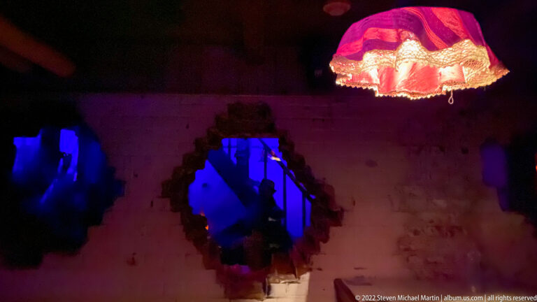 Dark damaged-room installation with jagged wall openings lit blue beneath a glowing red fabric lampshade.