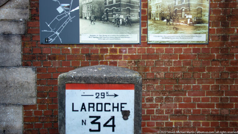 Concrete road marker reading LA ROCHE 34 beneath two historical photo panels on a brick wall.