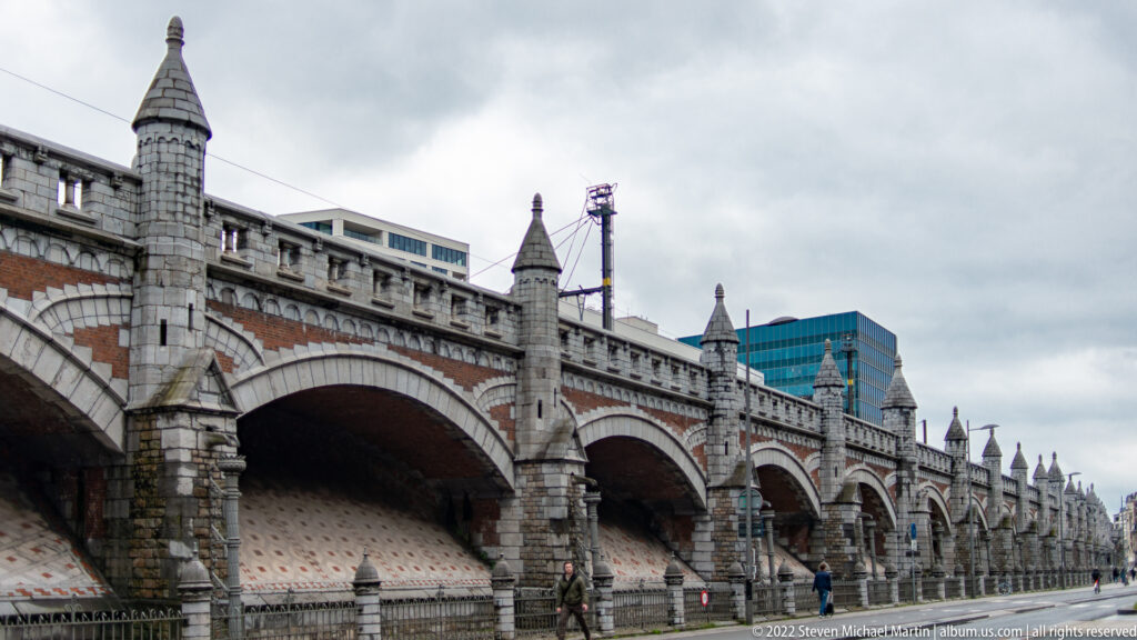 Historic brick railway viaduct with repeated stone-framed arches and pointed piers extending along a city street in Antwerp.