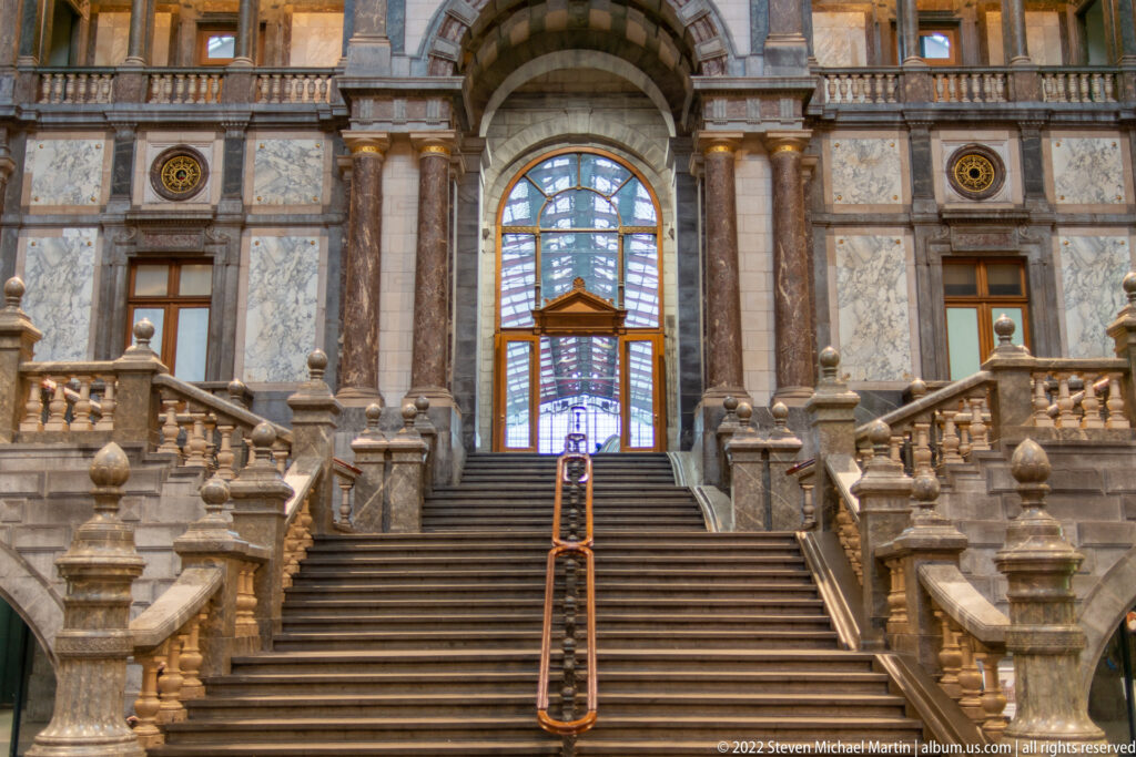 Symmetrical stone staircase with balustrades, columns, and a tall arched window inside Antwerp-Central Station.