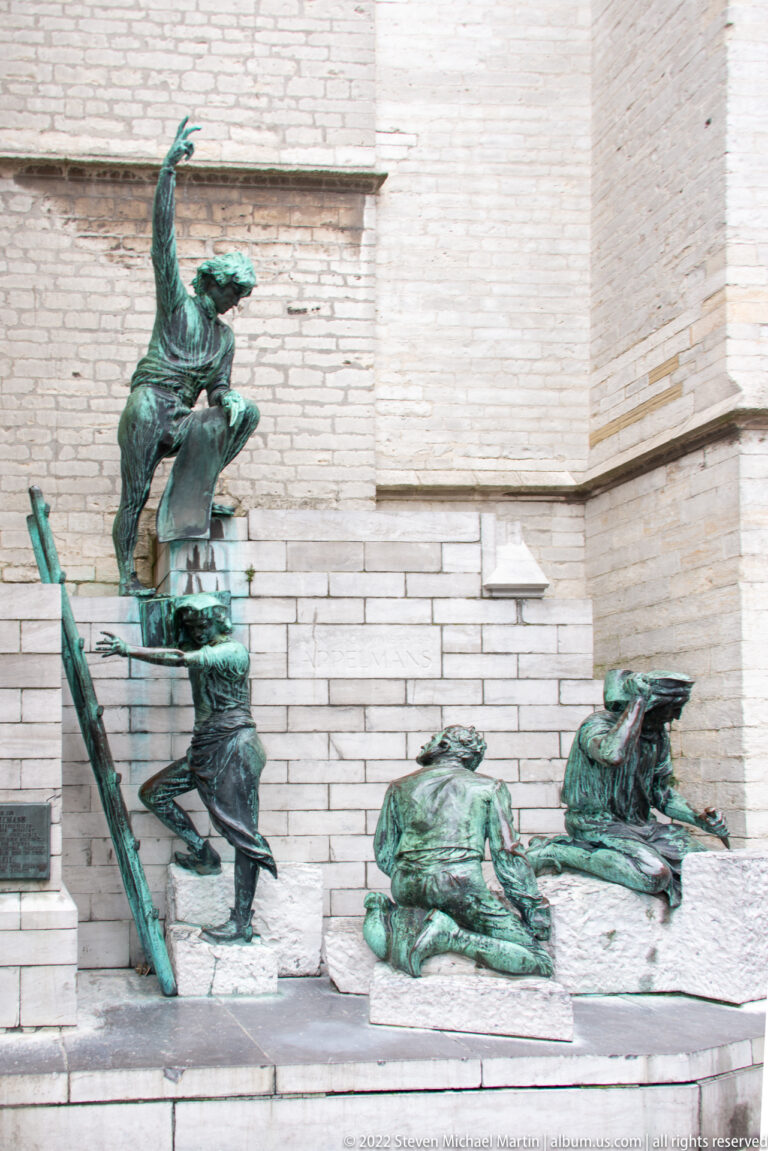 Cluster of green-patinated bronze human figures reclining, crouching, and rising beside a pale stone cathedral wall.