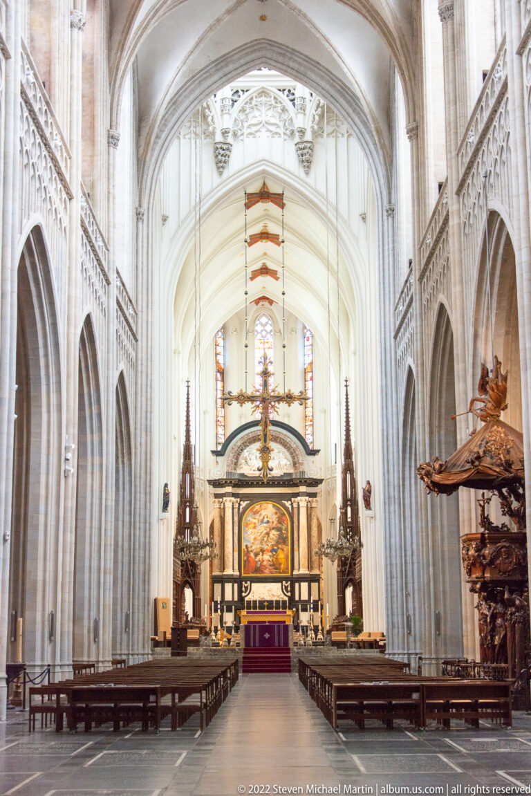 Cathedral sanctuary with monumental altarpiece, suspended cross, pale Gothic columns, vaulted ceiling, and rows of seating.