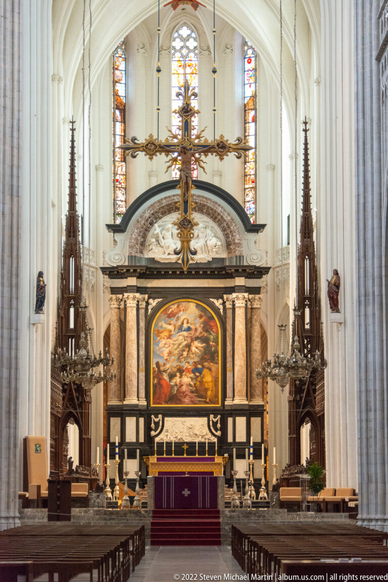 Monumental cathedral altar with large painted panel, dark architectural frame, suspended crucifix, and purple frontal cloth.