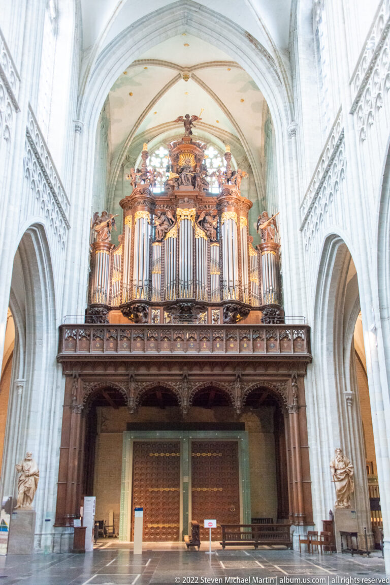 Large gilded and dark-wood pipe organ mounted above a gallery beneath the pale Gothic vaults of Antwerp Cathedral.