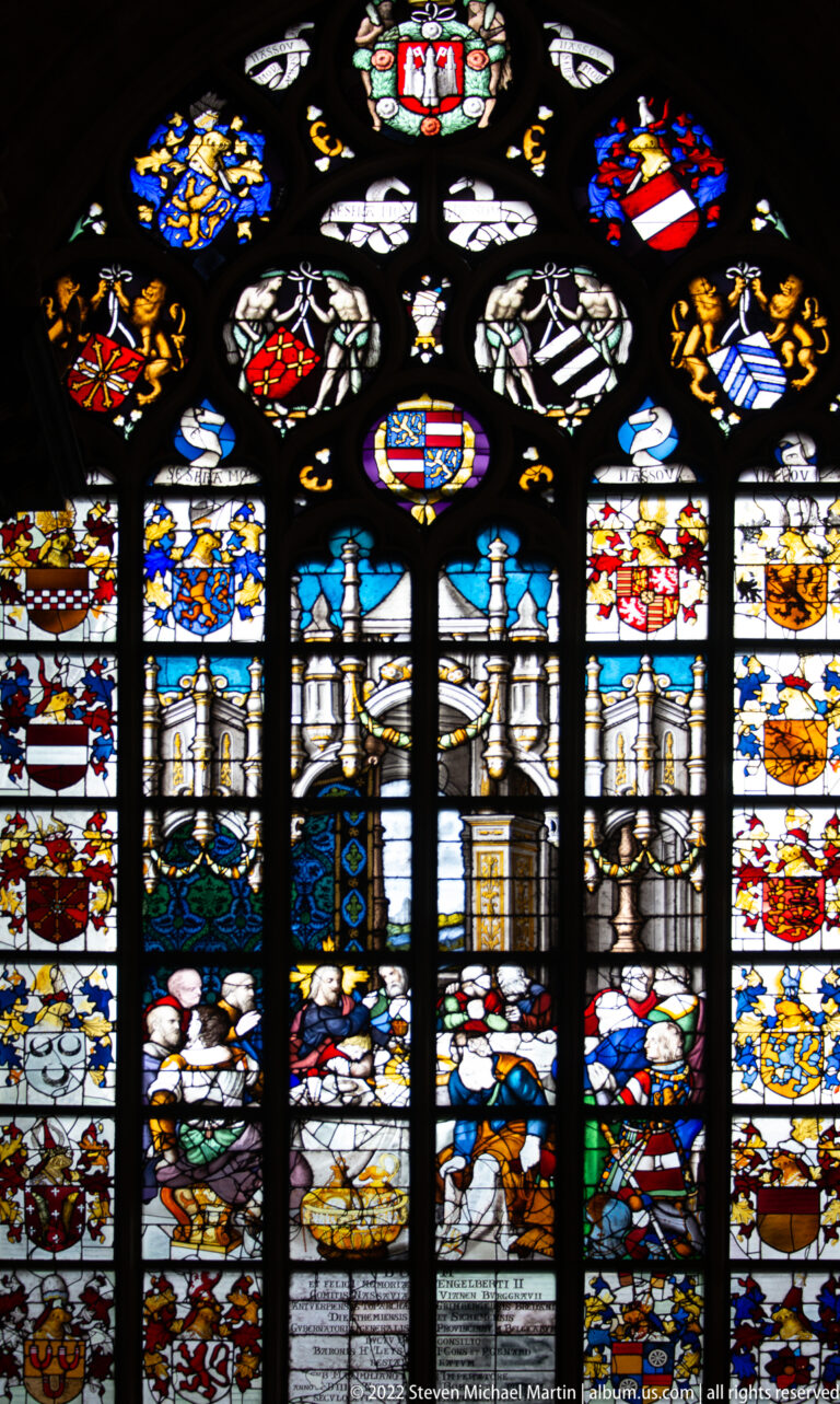 Large cathedral stained-glass window filled with heraldic shields, architectural motifs, figures, and dark lead tracery.