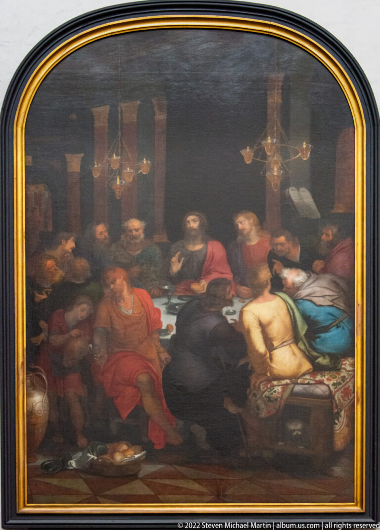 Large arched religious painting of figures gathered at a banquet table beneath chandeliers and columns, in a gilded frame.