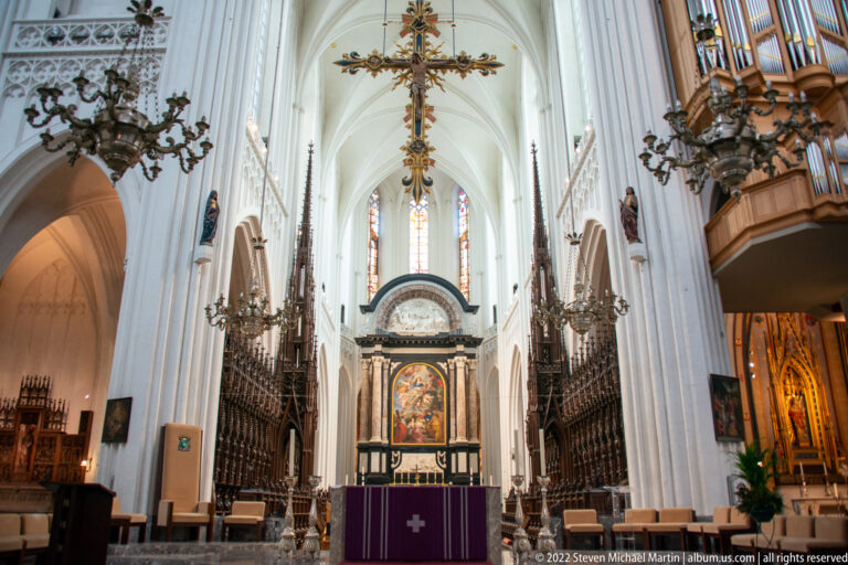 Symmetrical cathedral choir with dark carved stalls, central altar painting, suspended crucifix, Gothic vaulting, and organ pipes at right.