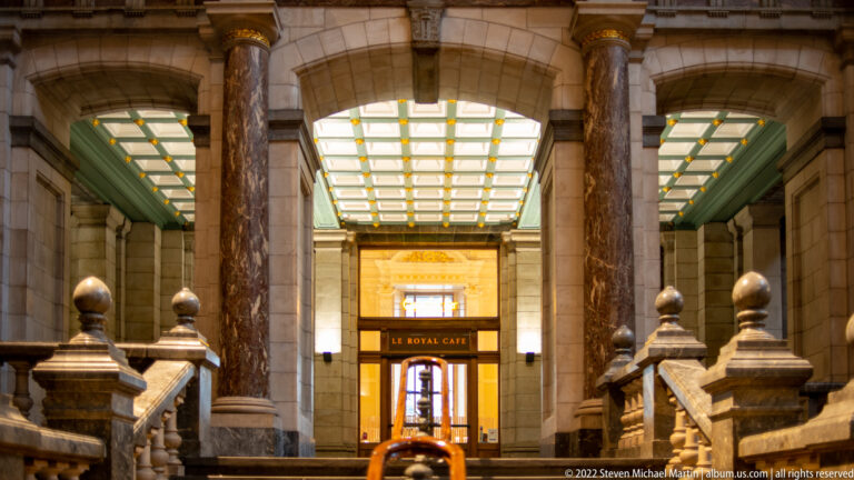 Le Royal Café entrance framed by polished columns, stone arches, bright ceiling panels, and stair finials.