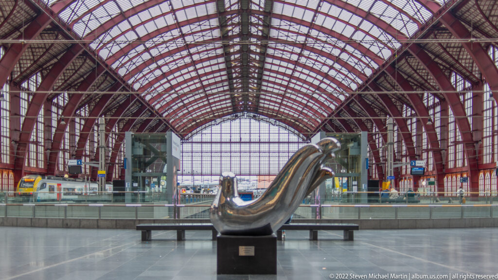Curving polished metal sculpture on a dark plinth beneath the red iron train shed of Antwerp-Central Station.