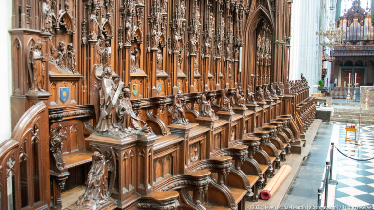 Long row of carved wooden choir stalls with saint figures, tracery, pinnacles, and small narrative reliefs inside Antwerp Cathedral.