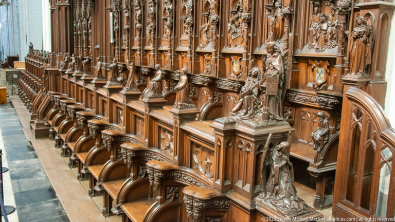 Close oblique view of carved cathedral choir stalls with sculpted saints, pointed canopies, armrests, and repeated wooden seats.