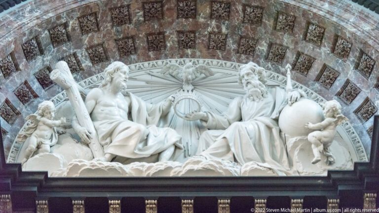 White marble allegorical relief with two reclining figures, putti, globe, cross-like staff, and Latin inscriptions beneath an arch.