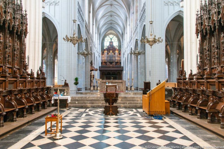 Symmetrical cathedral choir with carved stalls, black-and-white floor, Gothic vaults, chandeliers, distant organ, and stained glass.