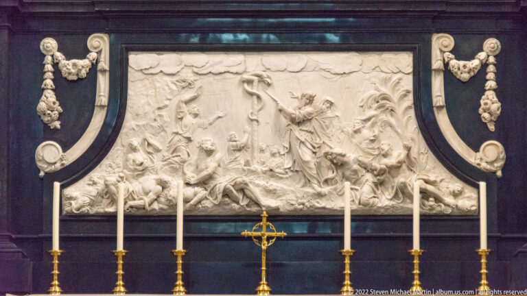 Wide white relief of a crowded religious scene above an altar lined with six tall candles.