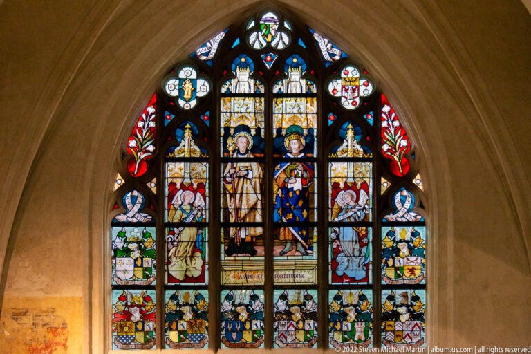 Pointed stained-glass window with standing figures, heraldic shields, bright colored panels, and dark lead tracery.