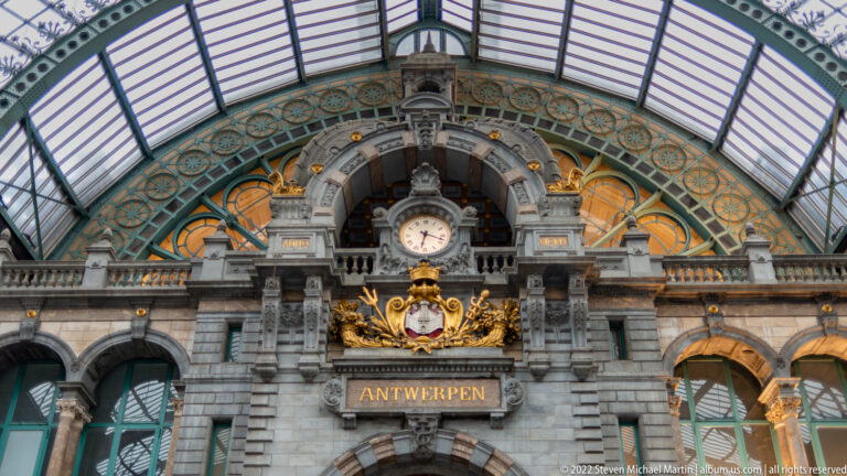Ornate Antwerp-Central Station facade with clock, gilded crest, and large ANTWERPEN sign beneath the glass roof.