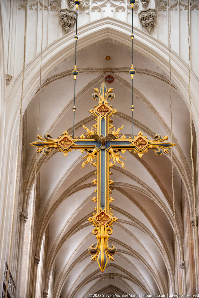 Large ornate gilded cross suspended beneath pale Gothic rib vaults inside Antwerp Cathedral.