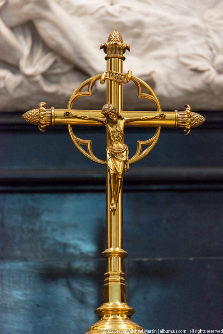 Gilded altar crucifix with a small figure of Christ, decorative finials, and a dark blue-black background.