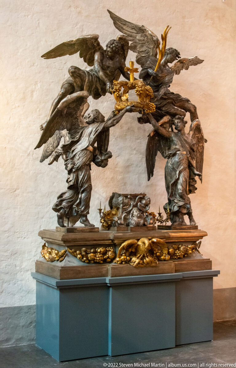 Bronze sculptural group with winged figures, kneeling figures, skull, and a gilded cross on a low base.