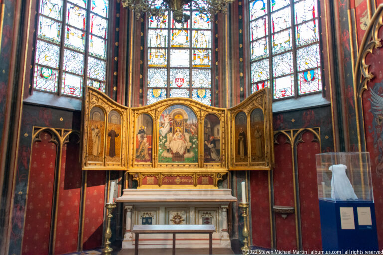 Small chapel altar with gilded multi-panel altarpiece, painted saints, stained-glass windows, and red wall decoration.