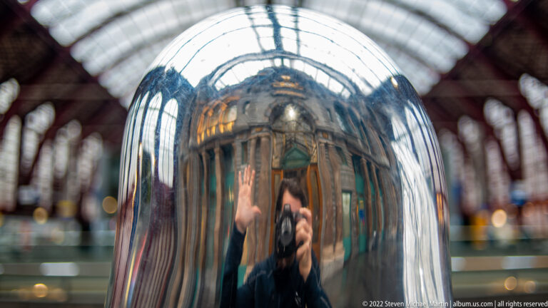 Close reflective metal surface showing the photographer and distorted arches of Antwerp-Central Station.