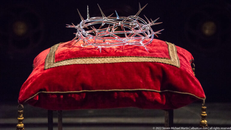 Large metal Crown of Thorns resting on a red velvet cushion with gold trim in a dark display case.