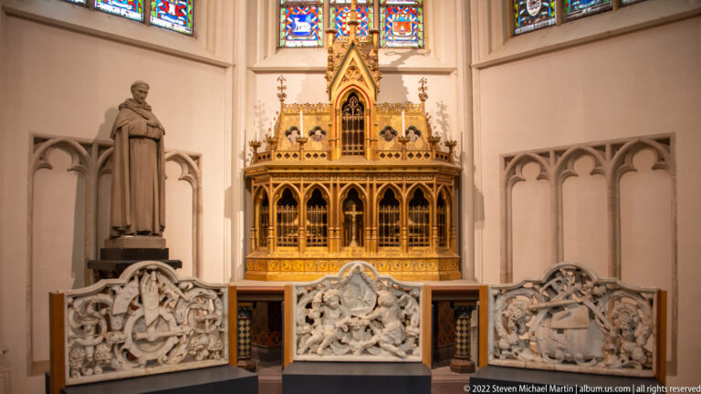 Gilded Gothic-style shrine beneath stained glass, with a standing saint and three pale carved relief panels.